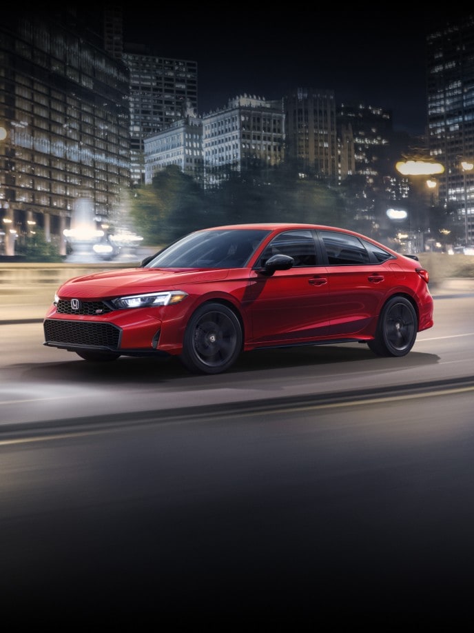 2026 Honda Civic Si Sedan in Rallye Red driving through the night time cityscape, with illuminated buildings in the background.