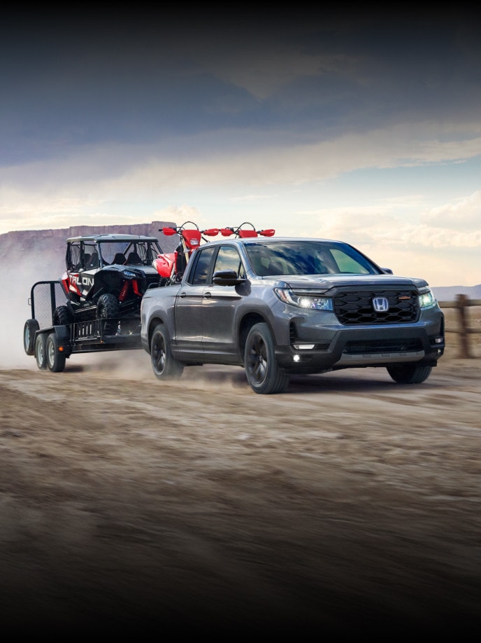 2026 Honda Ridgeline Trailsport in Modern Steel Metallic drives through a dirt field, carrying 2 dirt bikes in the flat bed and towing a Honda Talon on a trailer.